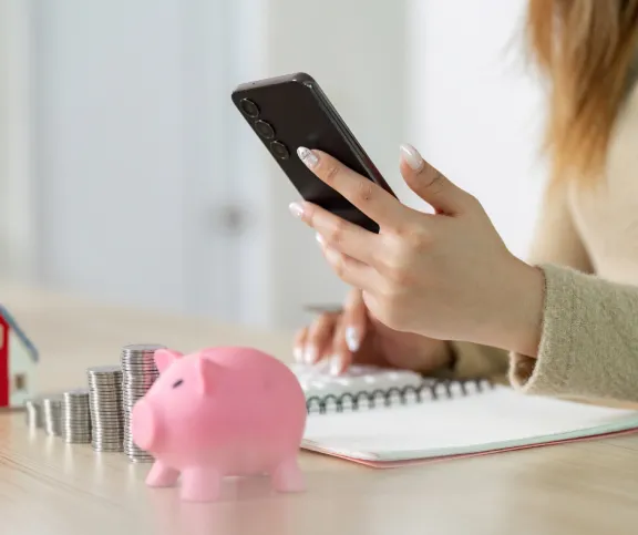 A woman holding her phone looking at her finances, piggy bank and coins in the foreground.